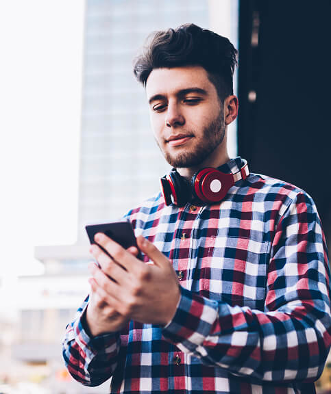 Young guy wearing headphones around neck and plaid shirt holding cell phone in both hands