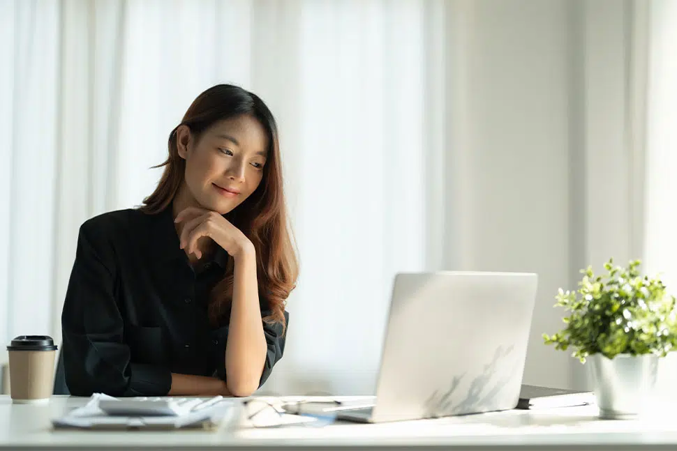 Asian woman in business clothes resting chin on hand while looking at desk with laptop, coffee and plant on surface.