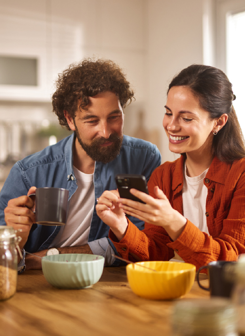 Man and woman sitting at kitchen table with bowls and coffee cup in hand looking at cell phone screen