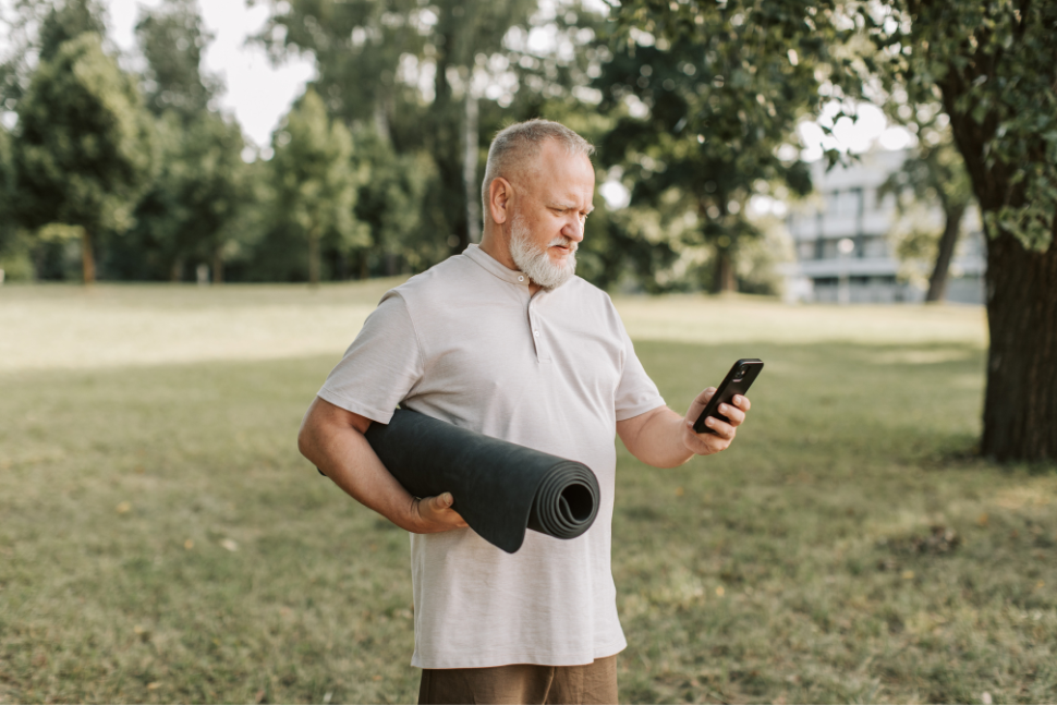 Older man in the park, carrying a rolled yoga mat and looking at his cell phone with a concerned look on his face