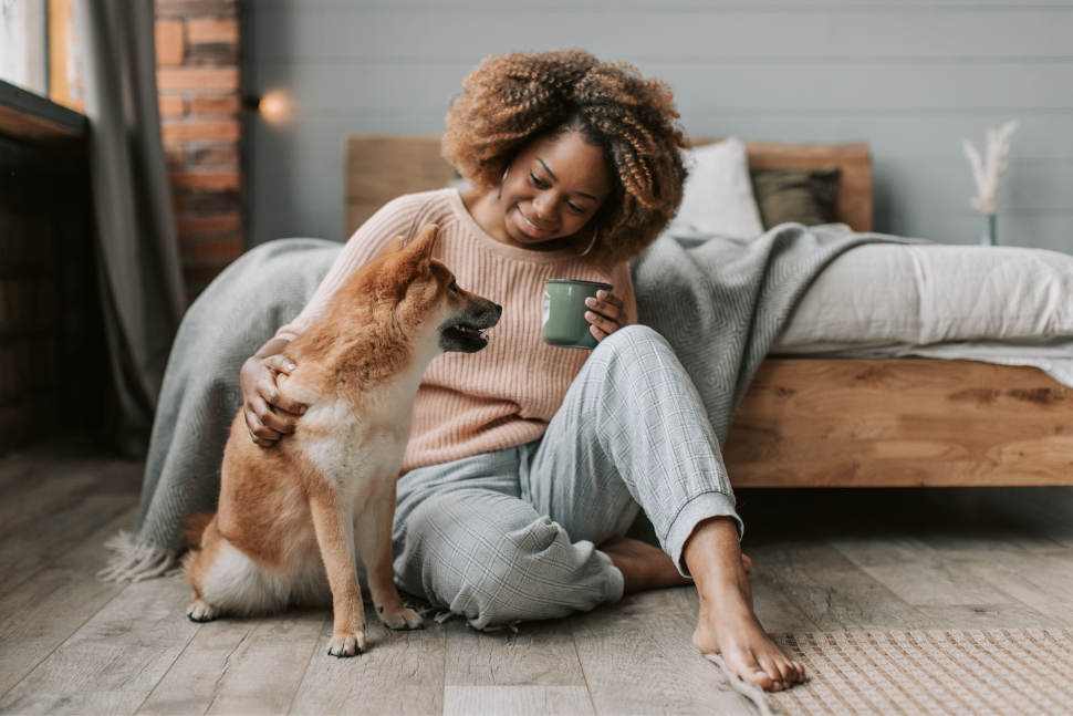 Image of an african american woman sitting on the floor, drinking from a mug and petting her dog