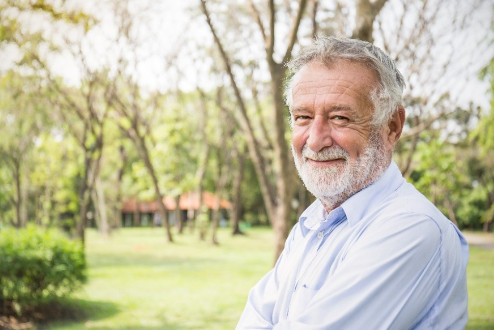 Image of a senior white gentleman wearing a light blue dress shirt and looking at the camera. The background is a park or yard.