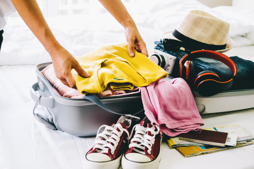 Image of an open suitcase. A set of hands is placing a folded shirt inside. There are other items including a pair of shoes, snorkel mask, hat, etc.